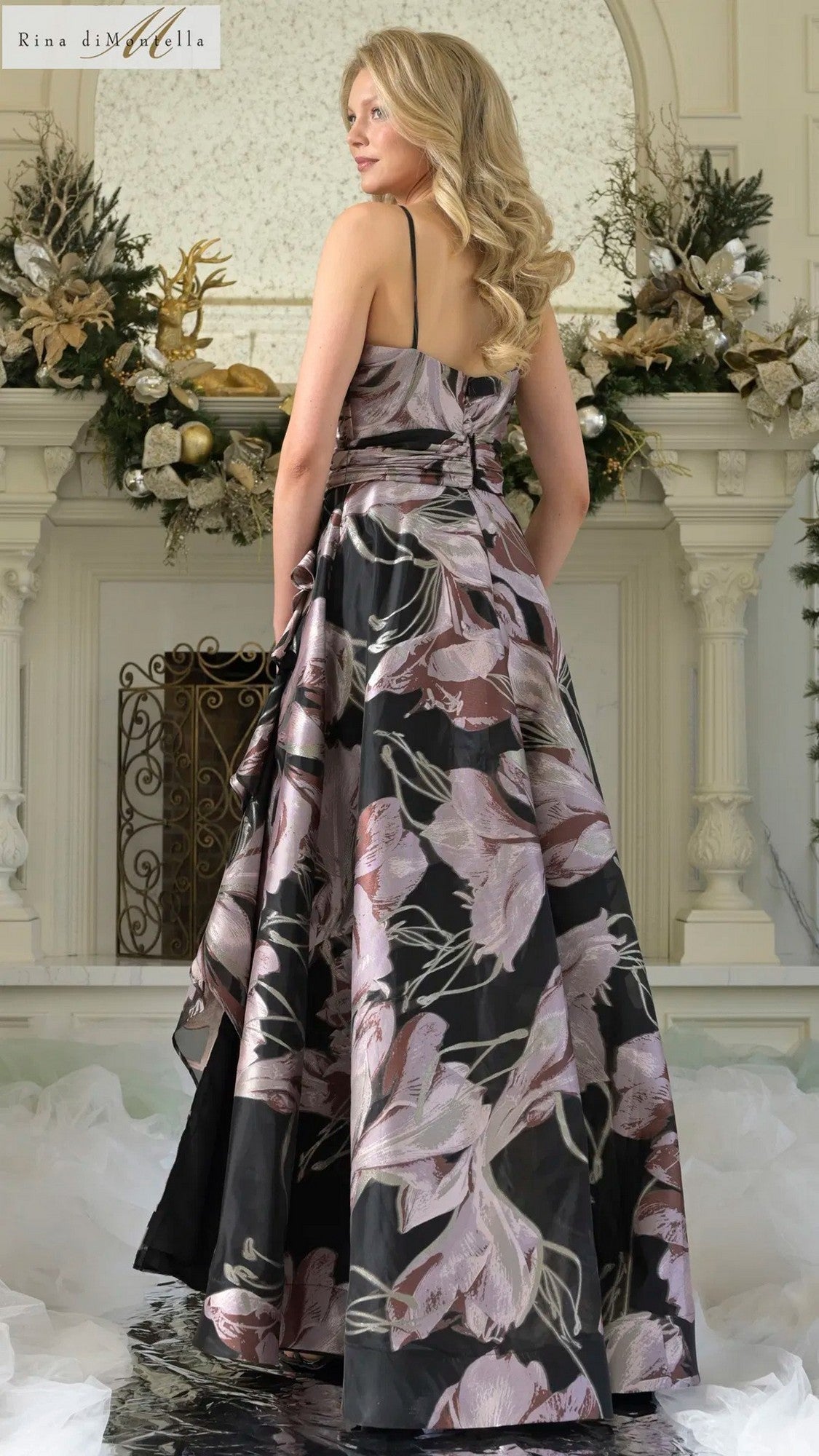 A woman in the Rina Di Montella RD3361 long formal dress stands before a festive fireplace with gold and green holiday accents, her elegant sleeveless floral jacquard gown enhancing the celebratory mood.